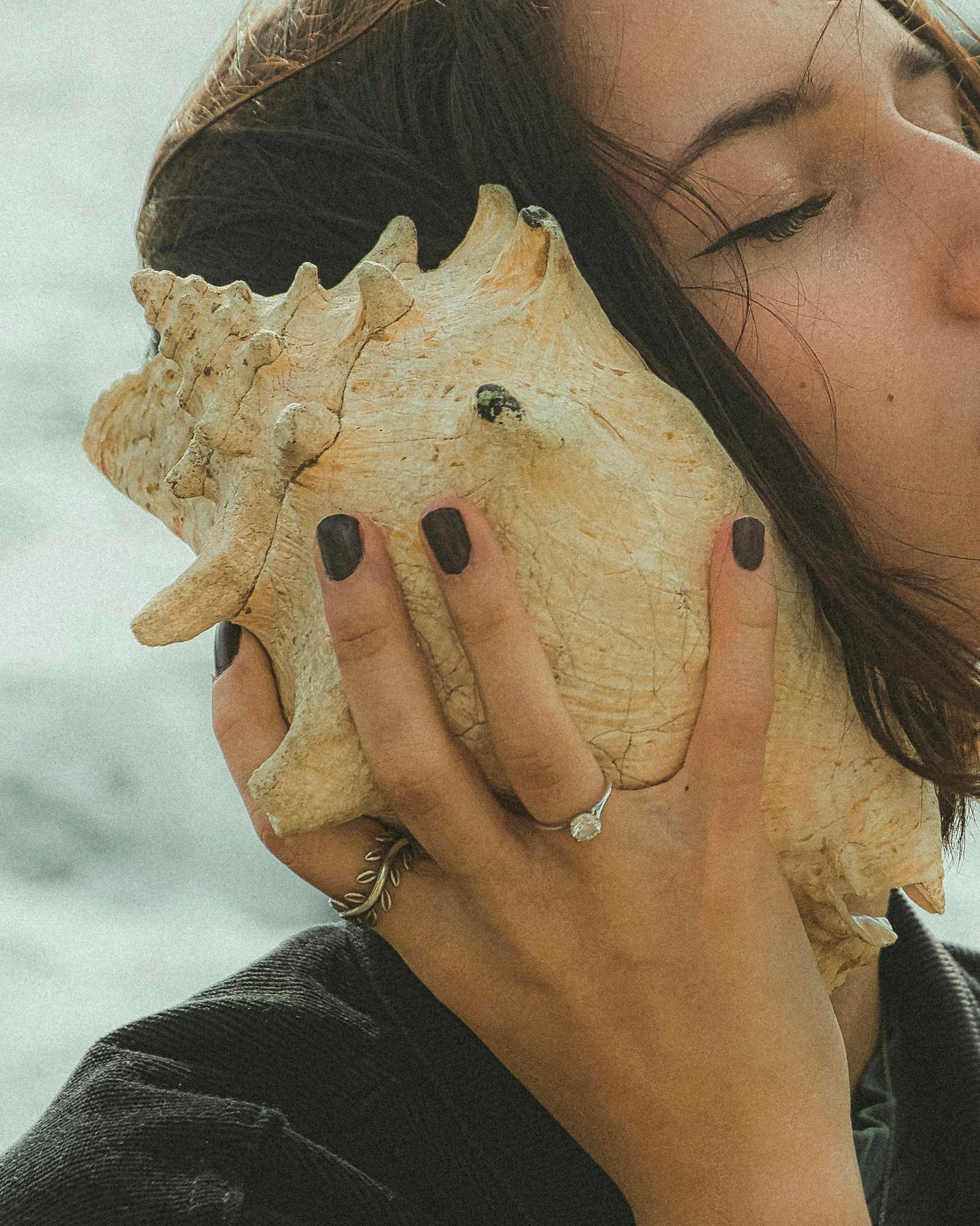 Woman with long hair holding a conch shell by her ear near the ocean.
